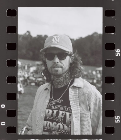 Monochrome portrait of a man with tattoos, wearing sunglasses and a baseball cap, posing in front of a blurred outdoor scene.