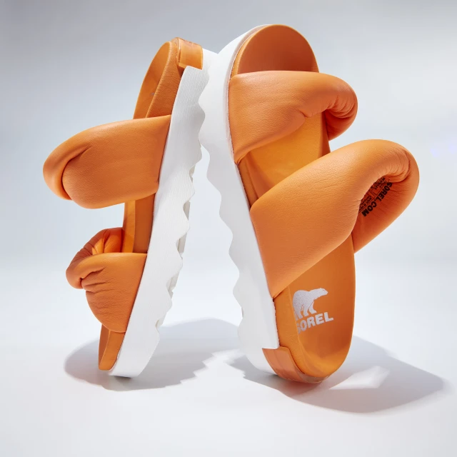 Orange SOREL sandals featuring puffy straps and a textured white sole, presented on a gradient white background in a studio product shot.