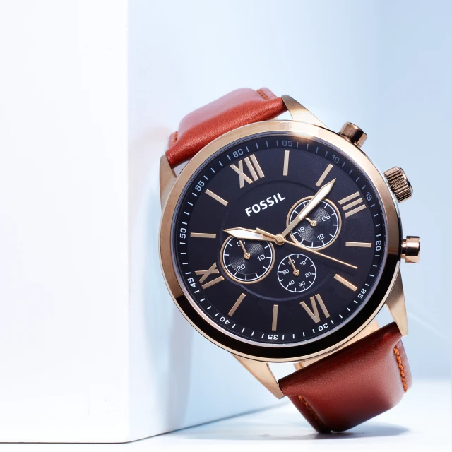 Fossil men's watch with a brown leather strap and a black face with Roman numerals sits on a white surface in a studio setting.