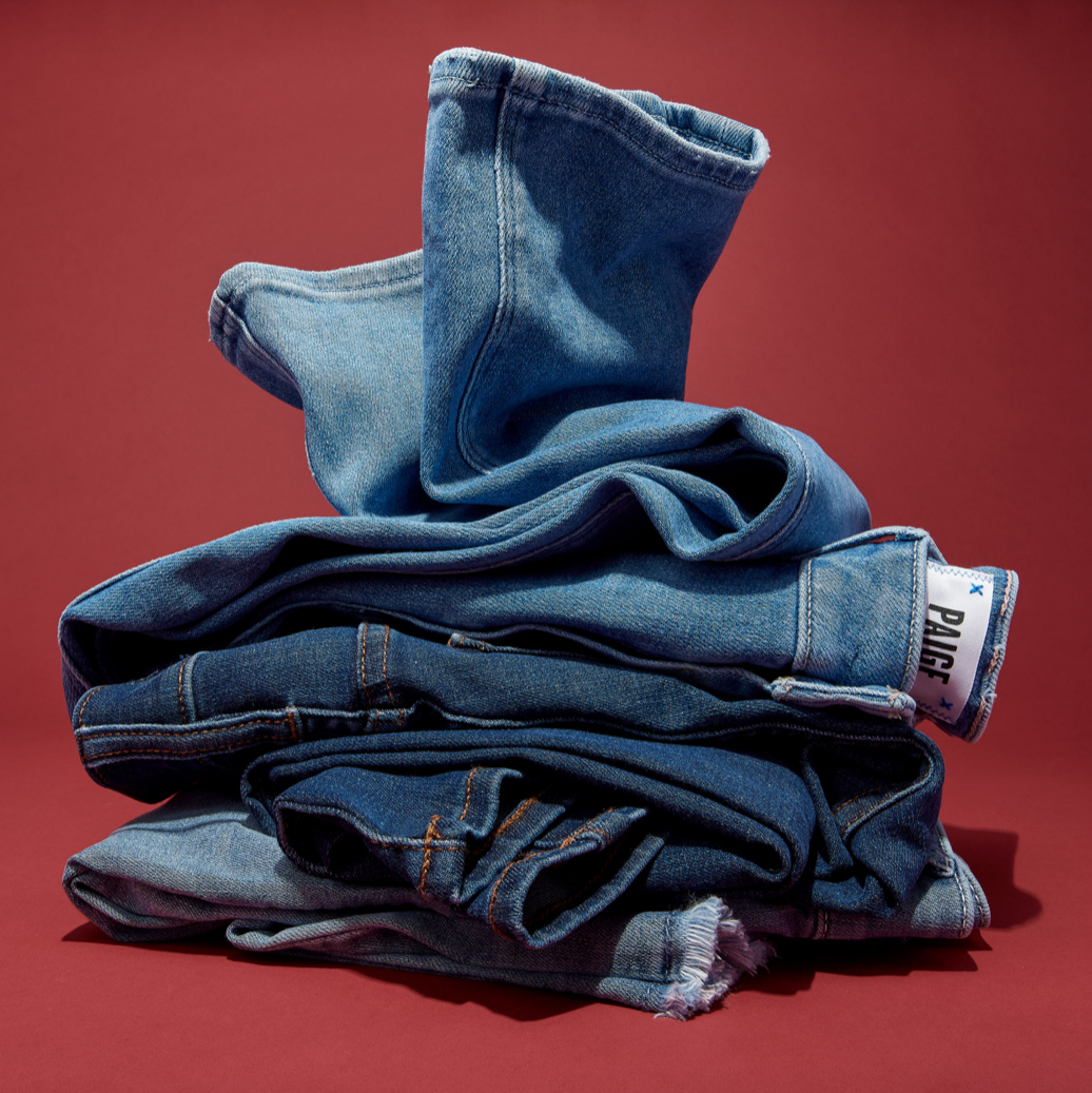 Stack of folded and partially folded blue denim jeans on a plain red background with soft studio lighting; label of jeans is visible.