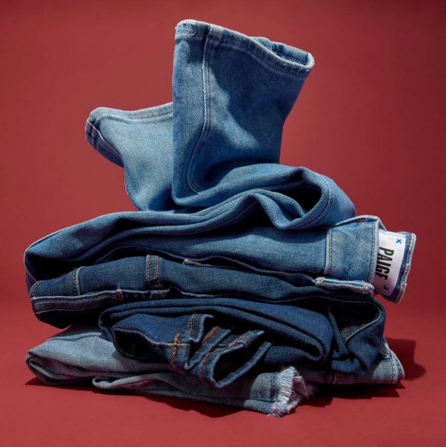 Stack of folded and partially folded blue denim jeans on a plain red background with soft studio lighting; label of jeans is visible.