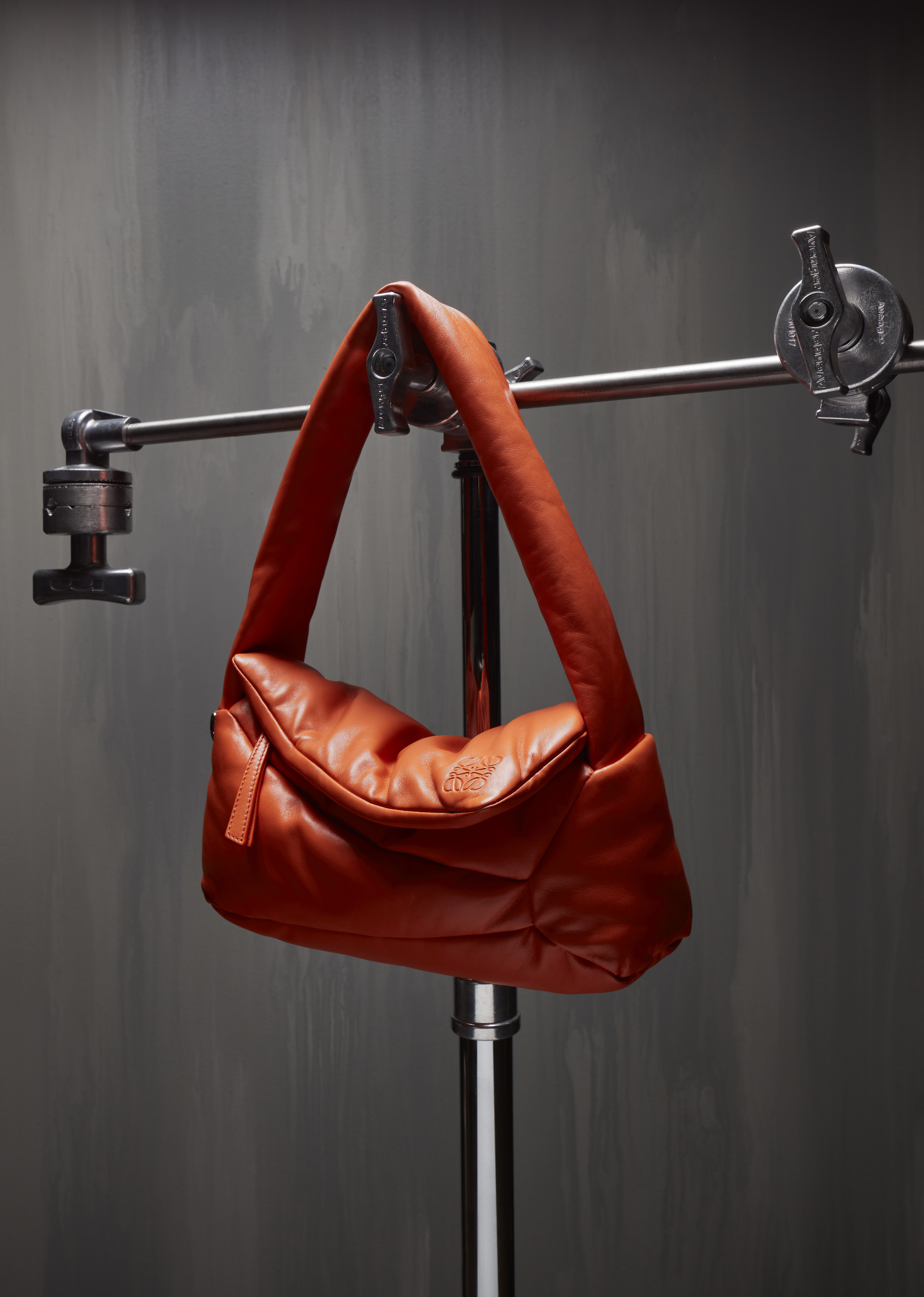 Studio shot of a vibrant orange leather handbag on a silver stand against a textured gray backdrop, fashion accessory, product photography.