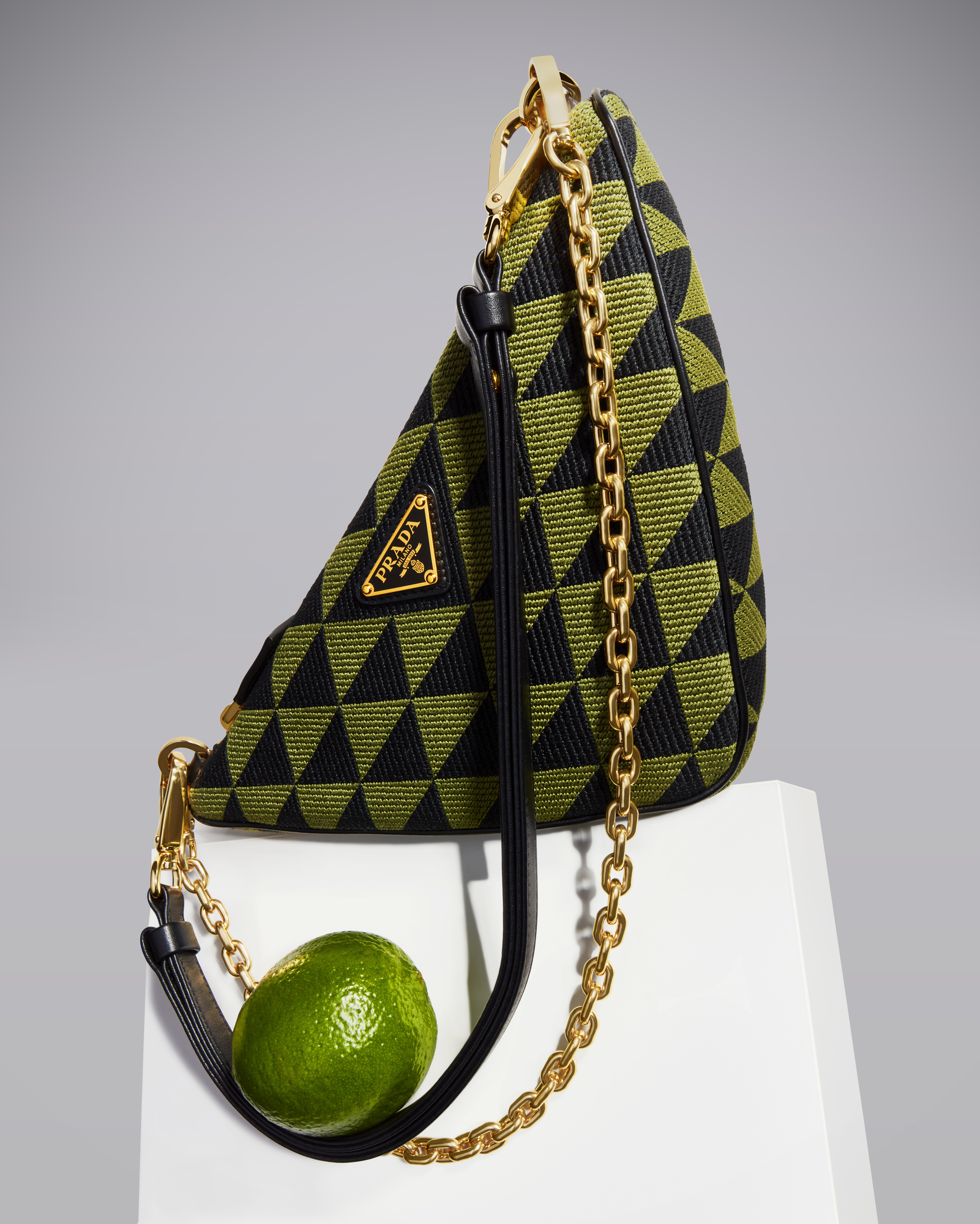 Prada triangle bag in black and olive green with geometric pattern, gold chain strap, leather handle, and lime fruit on white surface.