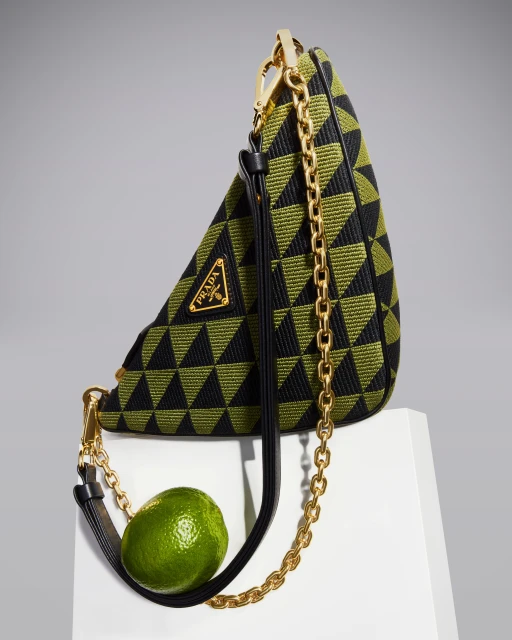 Prada triangle bag in black and olive green with geometric pattern, gold chain strap, leather handle, and lime fruit on white surface.