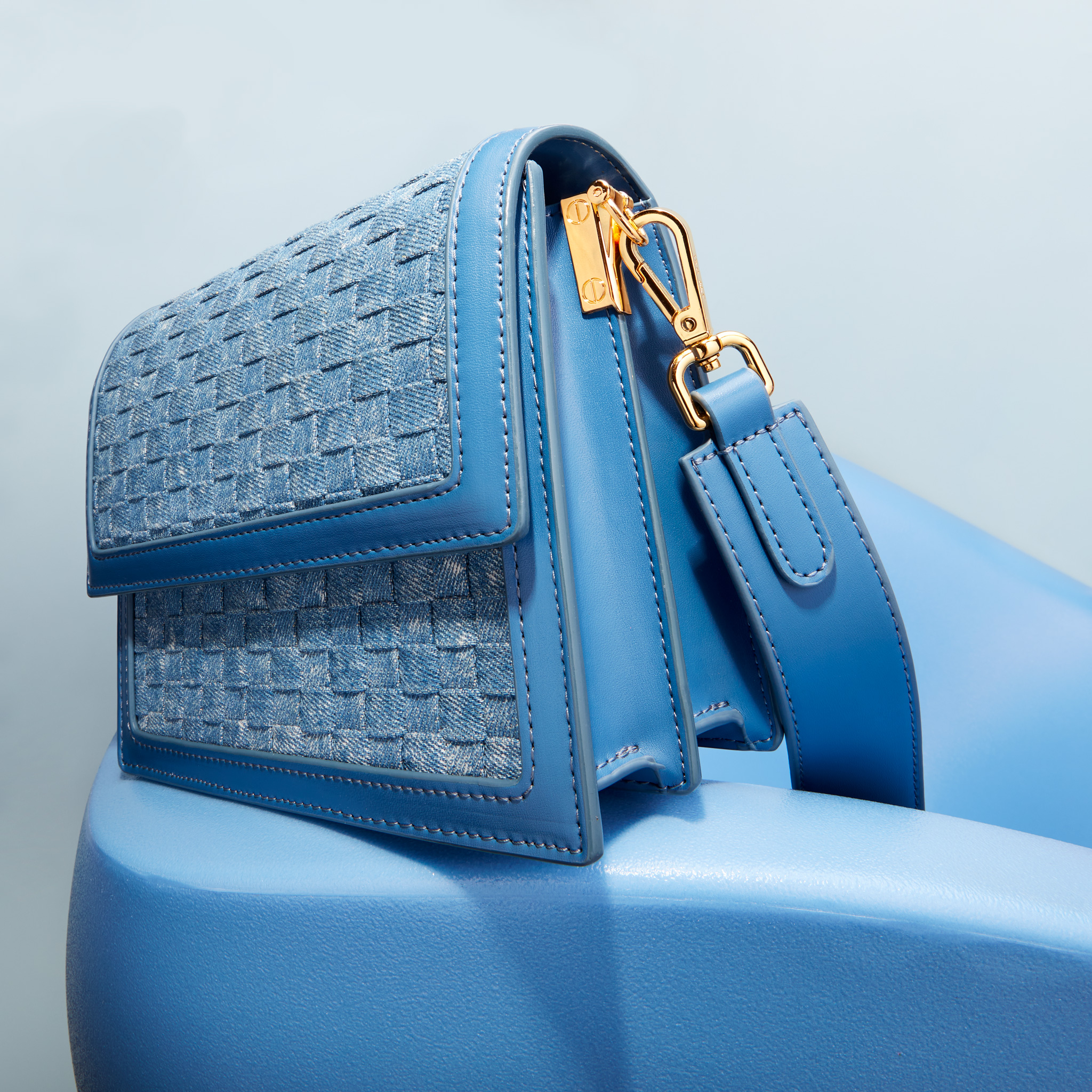 Close-up of a blue leather handbag with a woven flap and a gold clasp, resting on a blue surface in a minimalist studio setup.