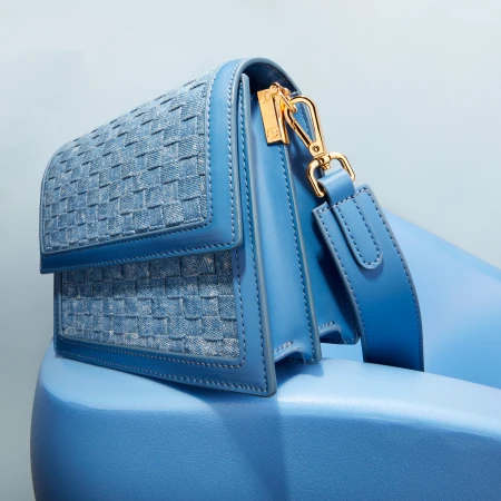 Close-up of a blue leather handbag with a woven flap and a gold clasp, resting on a blue surface in a minimalist studio setup.