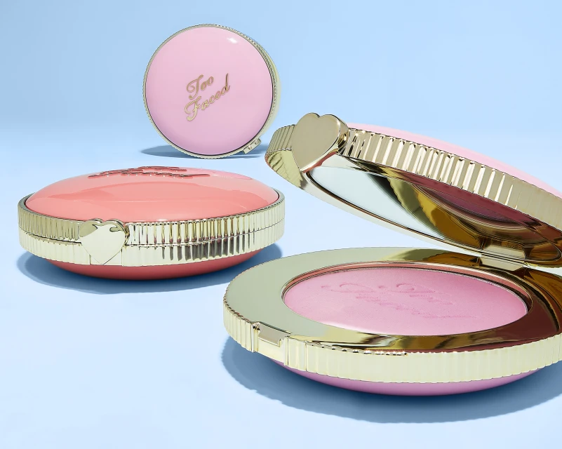 Three Too Faced cosmetic compacts in pink and gold on a light blue background; one is open, revealing pink powder inside.
