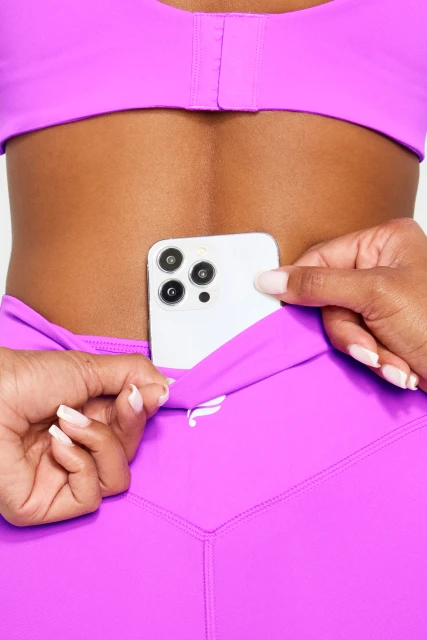 Close-up of a woman in a pink sports bra and leggings placing a white cell phone into the back pocket of her leggings.