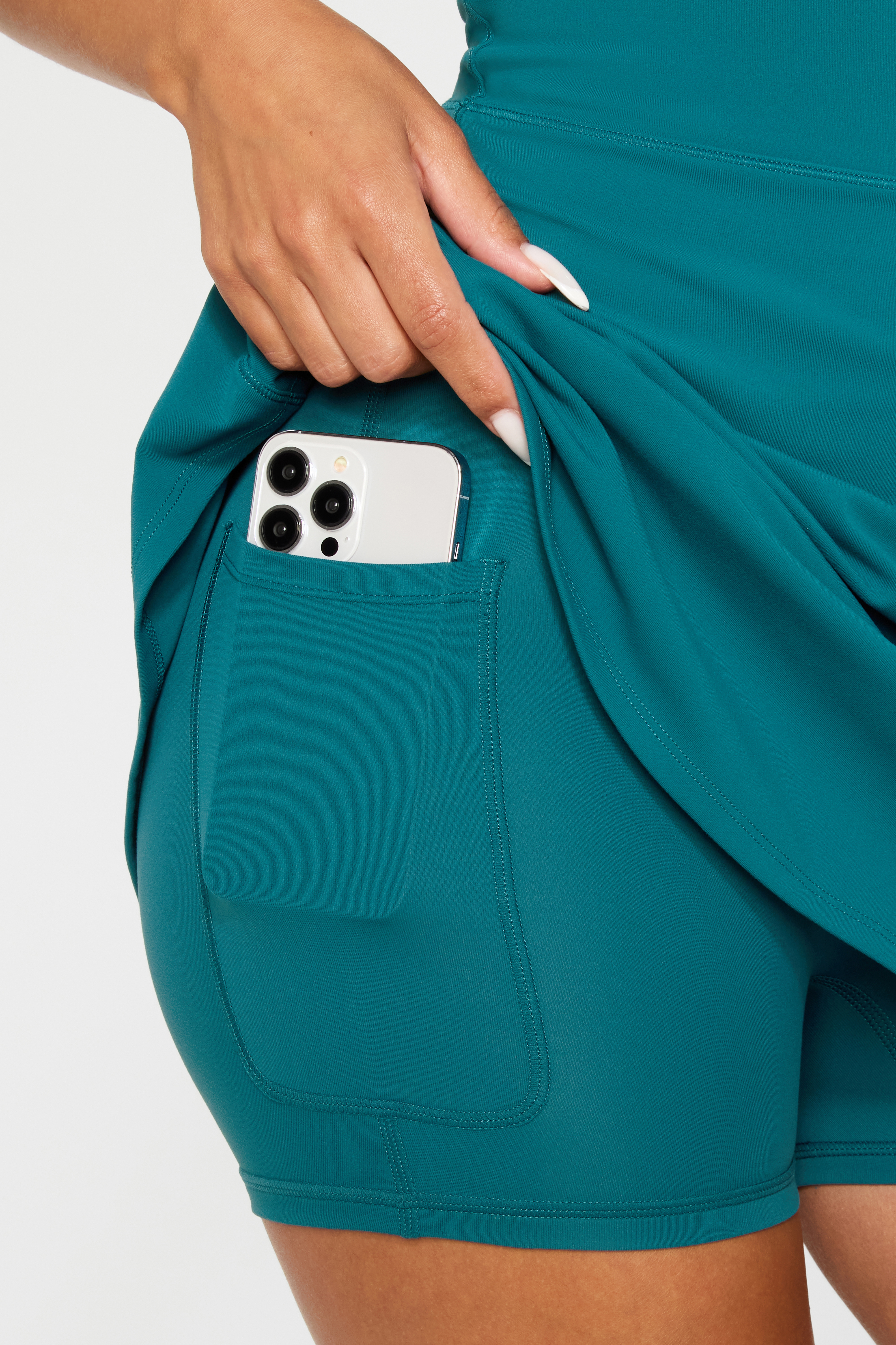 Woman wearing a teal athletic skirt with shorts and a side pocket holding a cell phone against a white background, studio shot.