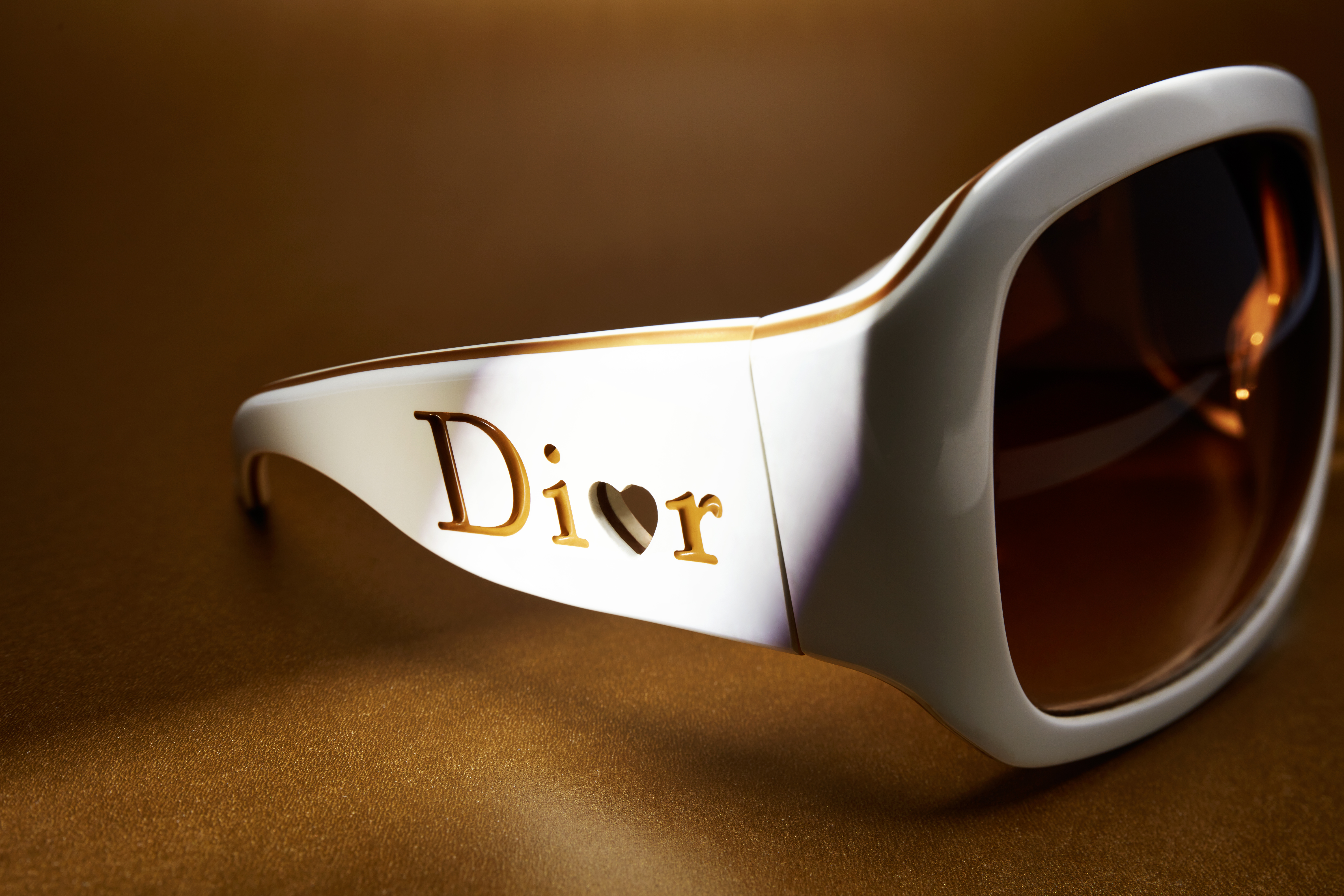 Close-up studio shot of white sunglasses with brown lenses. 'Dior' logo on the side, golden background, and stylish eye protection.