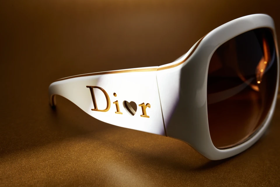 Close-up studio shot of white sunglasses with brown lenses. 'Dior' logo on the side, golden background, and stylish eye protection.