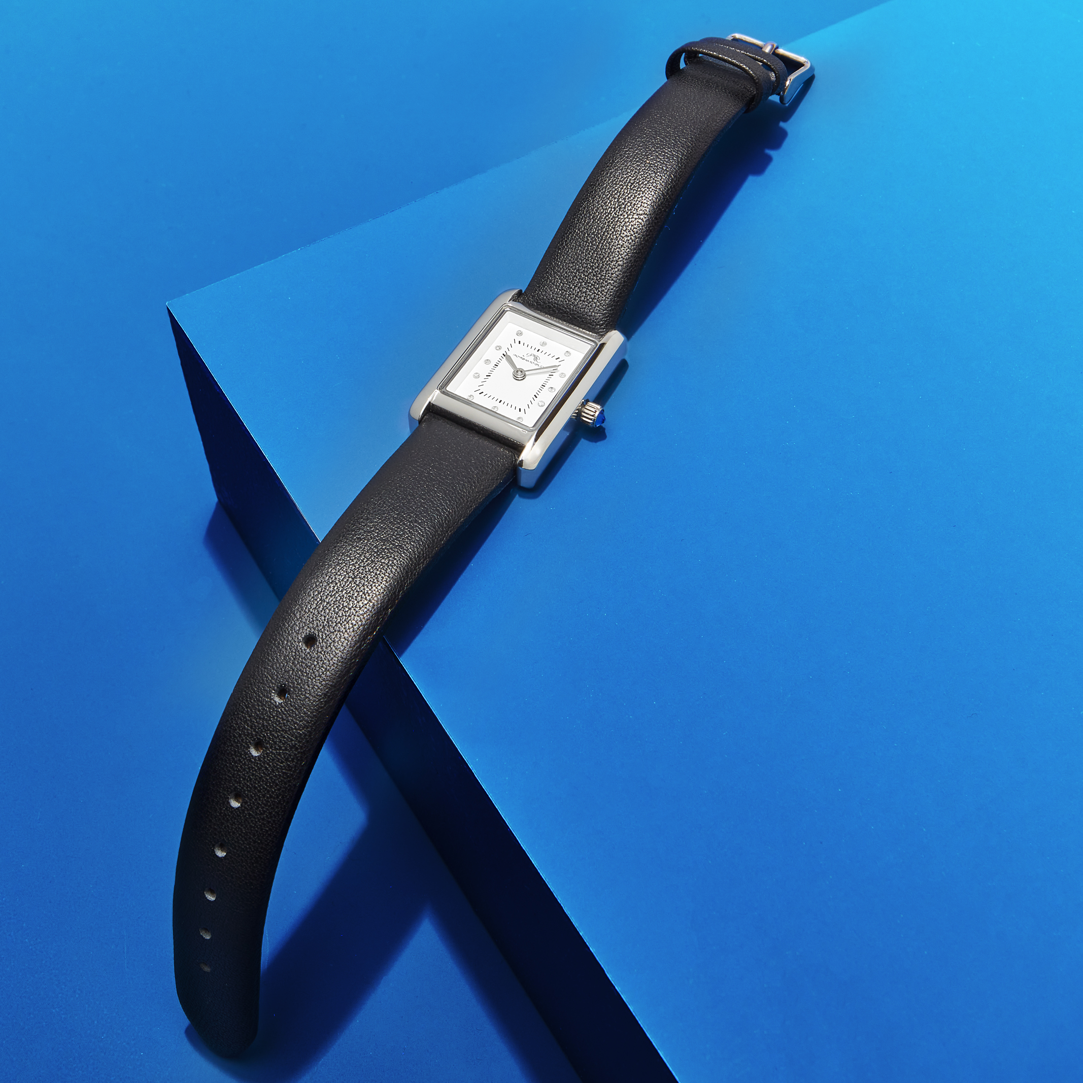 Elegant wristwatch with a silver rectangular face and black leather band, displayed on a vibrant blue geometric surface in studio lighting.