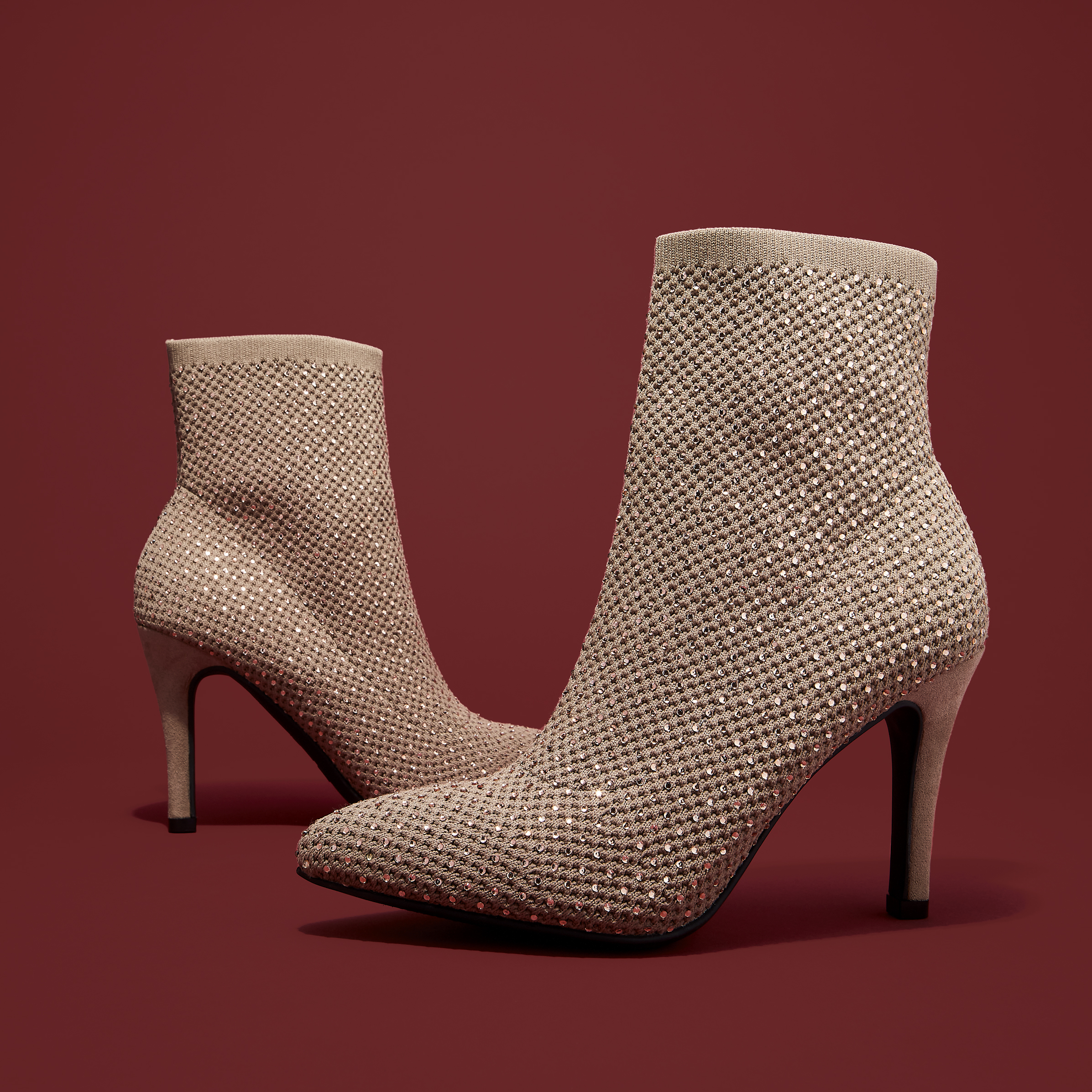 Beige sock-style high heel ankle boots embellished with rhinestones on a red background in a studio setting. Fashion footwear product shot.