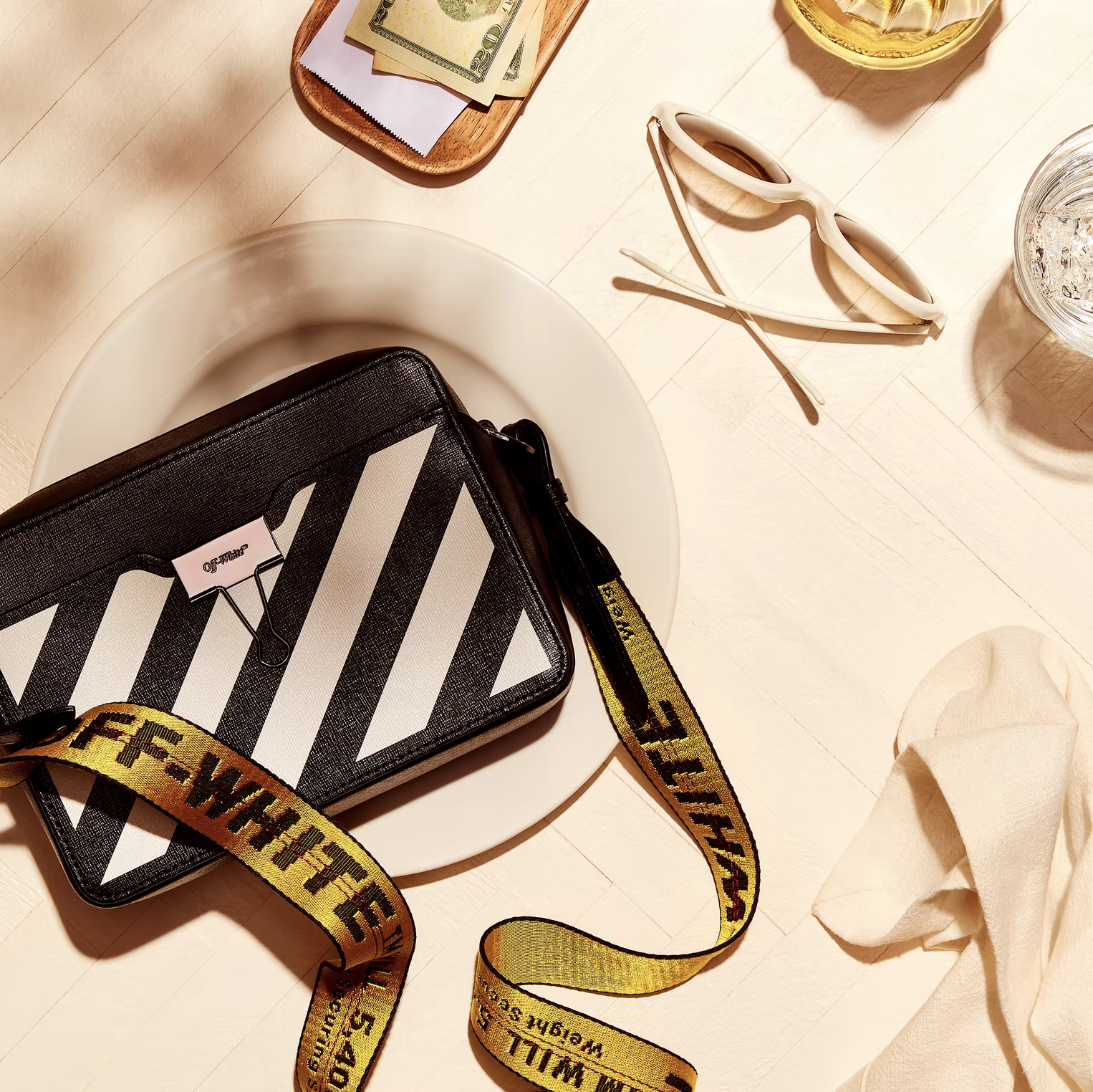 Overhead shot of a black and white Off-White handbag, sunglasses, money, and a glass of water on a white surface. 