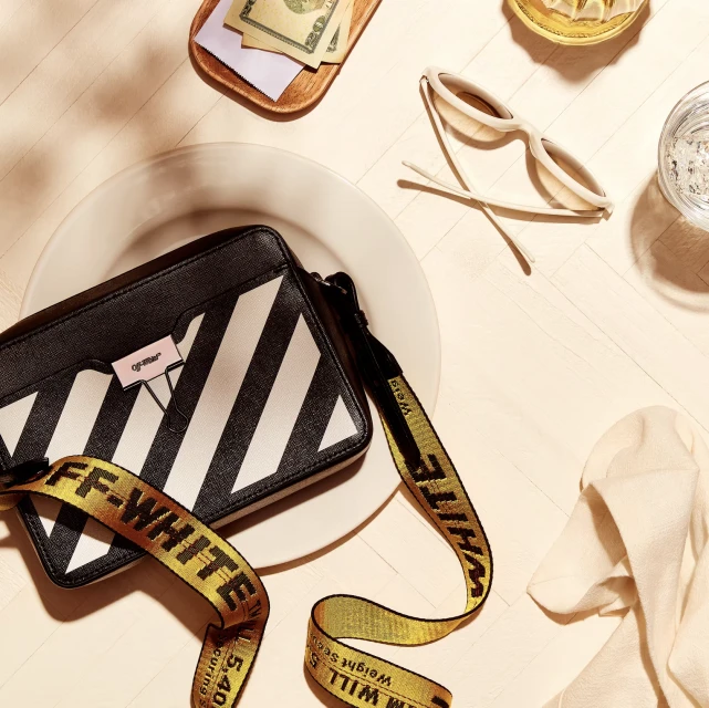 Overhead shot of a black and white Off-White handbag, sunglasses, money, and a glass of water on a white surface. 