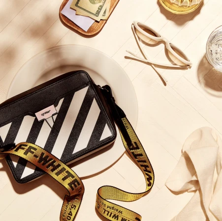 Overhead shot of a black and white Off-White handbag, sunglasses, money, and a glass of water on a white surface. 