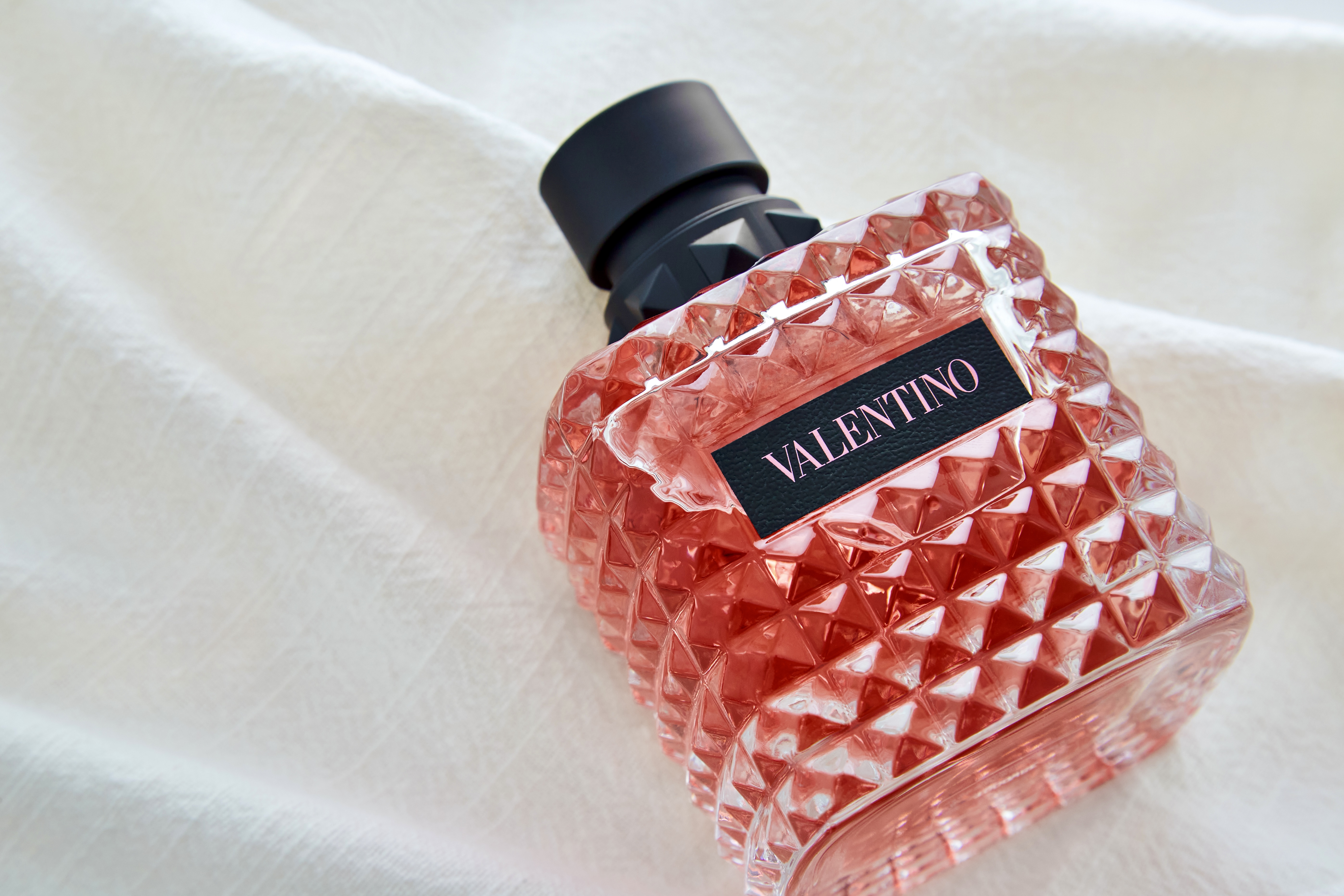 Close-up of a faceted, pink Valentino perfume bottle with a black cap and label, resting on a textured, off-white fabric backdrop.