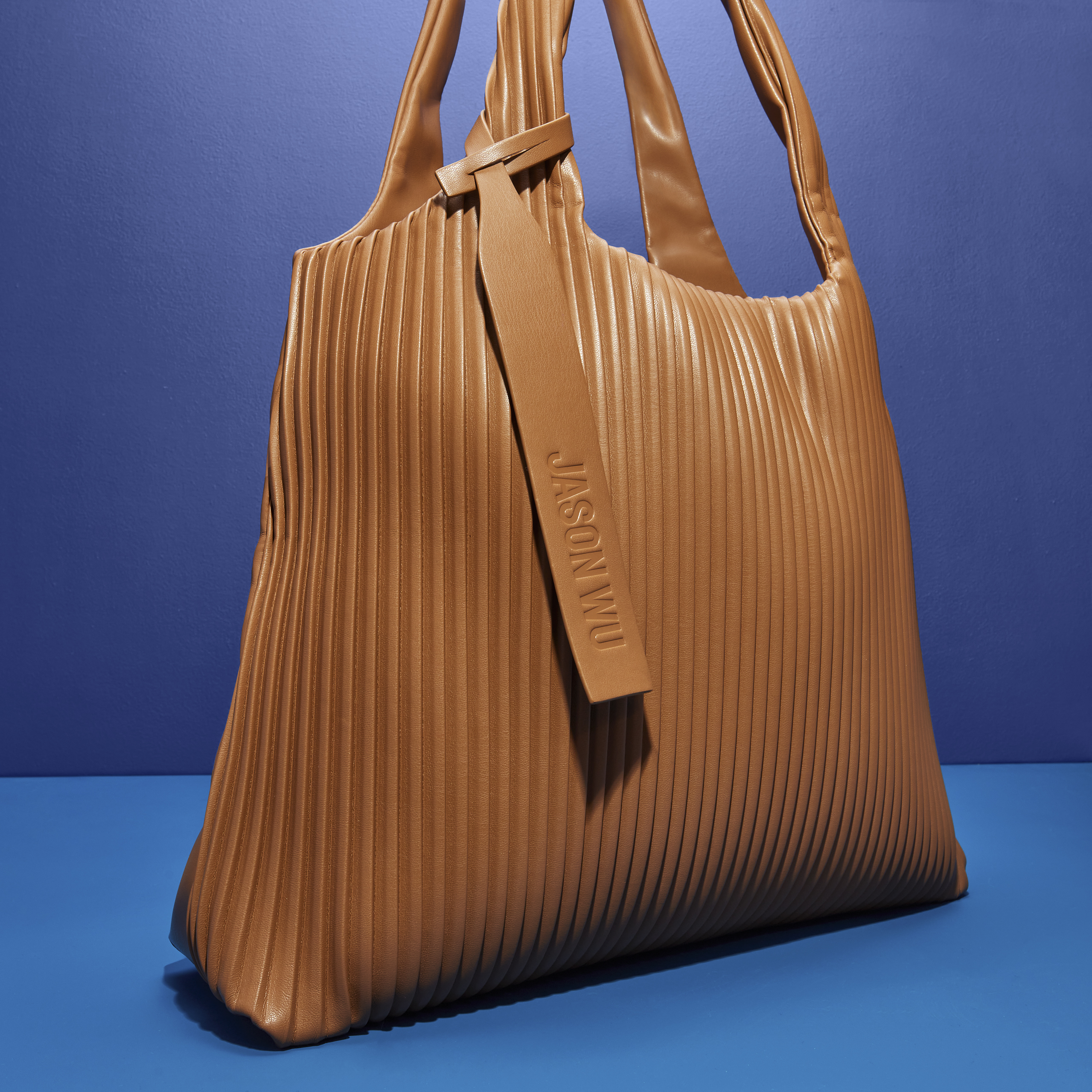 A brown pleated tote bag by Jason Wu is isolated against a blue background in a product shot showcasing its texture and design.