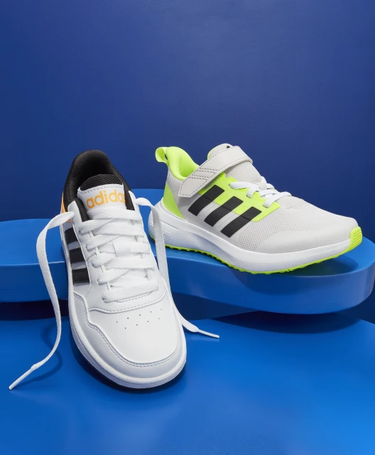 Two Adidas shoes with black stripes stand on a blue surface with a matching blue background. One shoe is white with black accents.