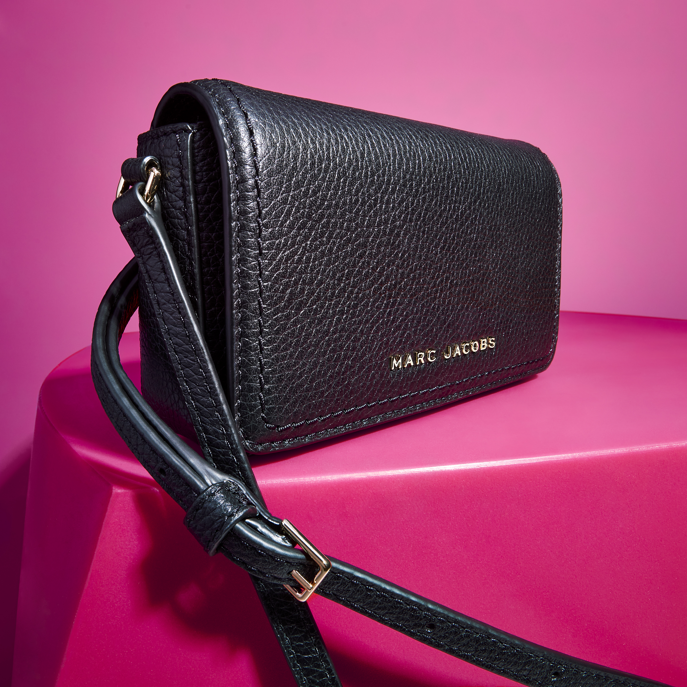 Black Marc Jacobs purse with a crossbody strap sits on a hot pink platform in a studio setting, showcasing its pebbled leather texture.