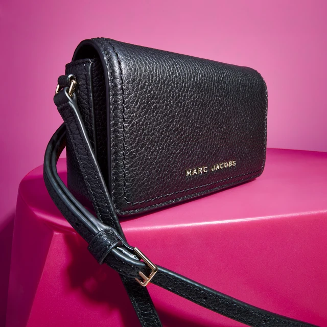 Black Marc Jacobs purse with a crossbody strap sits on a hot pink platform in a studio setting, showcasing its pebbled leather texture.