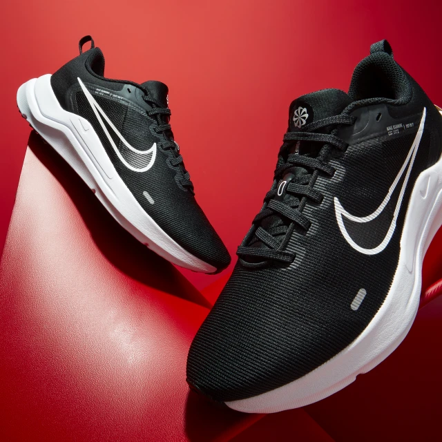 Two black Nike Downshifter 12 running shoes with white swooshes on a red studio backdrop. Athletic footwear product shot.