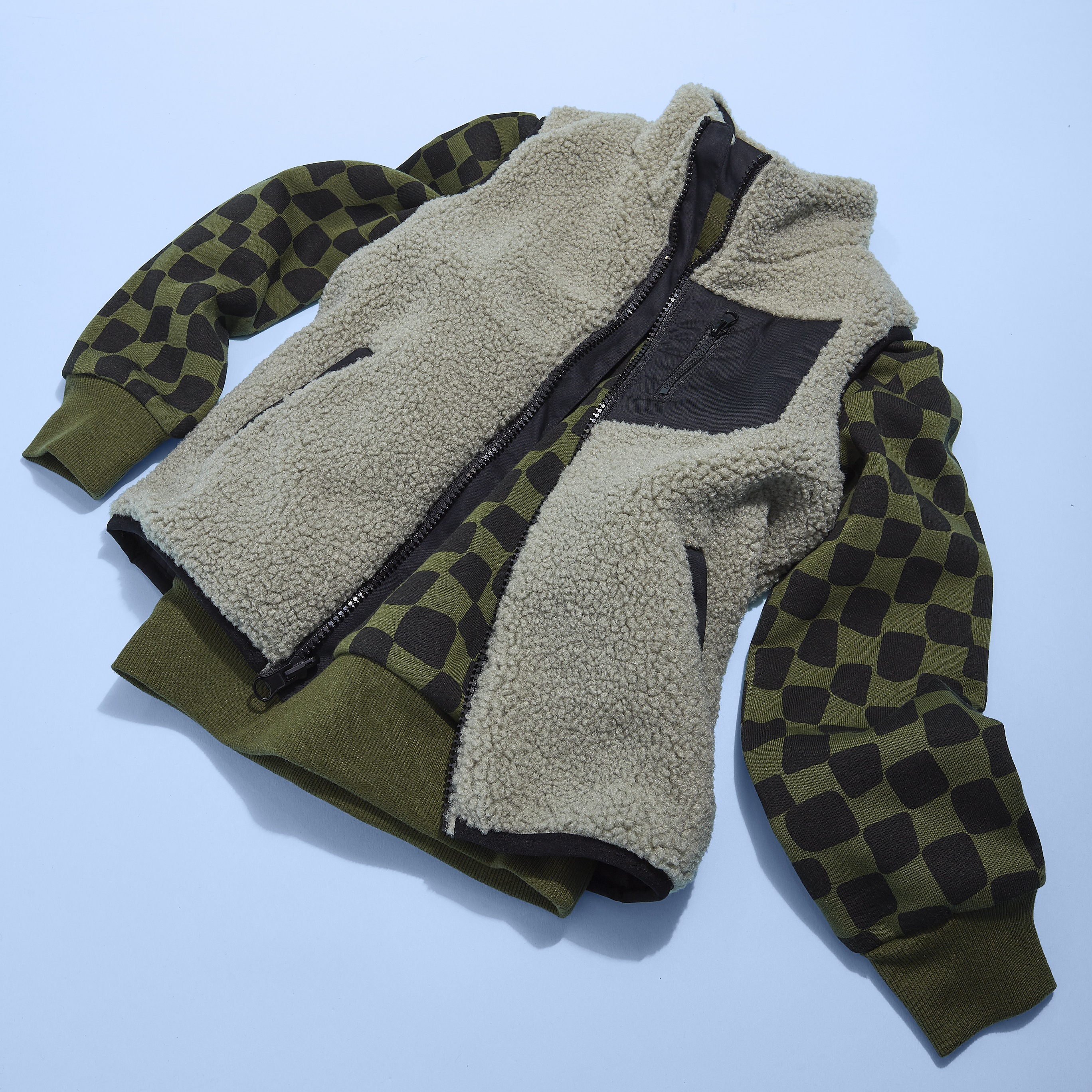 Olive green fleece vest with a checkered-sleeved jacket laid flat on a light blue surface in a studio setting with overhead lighting.