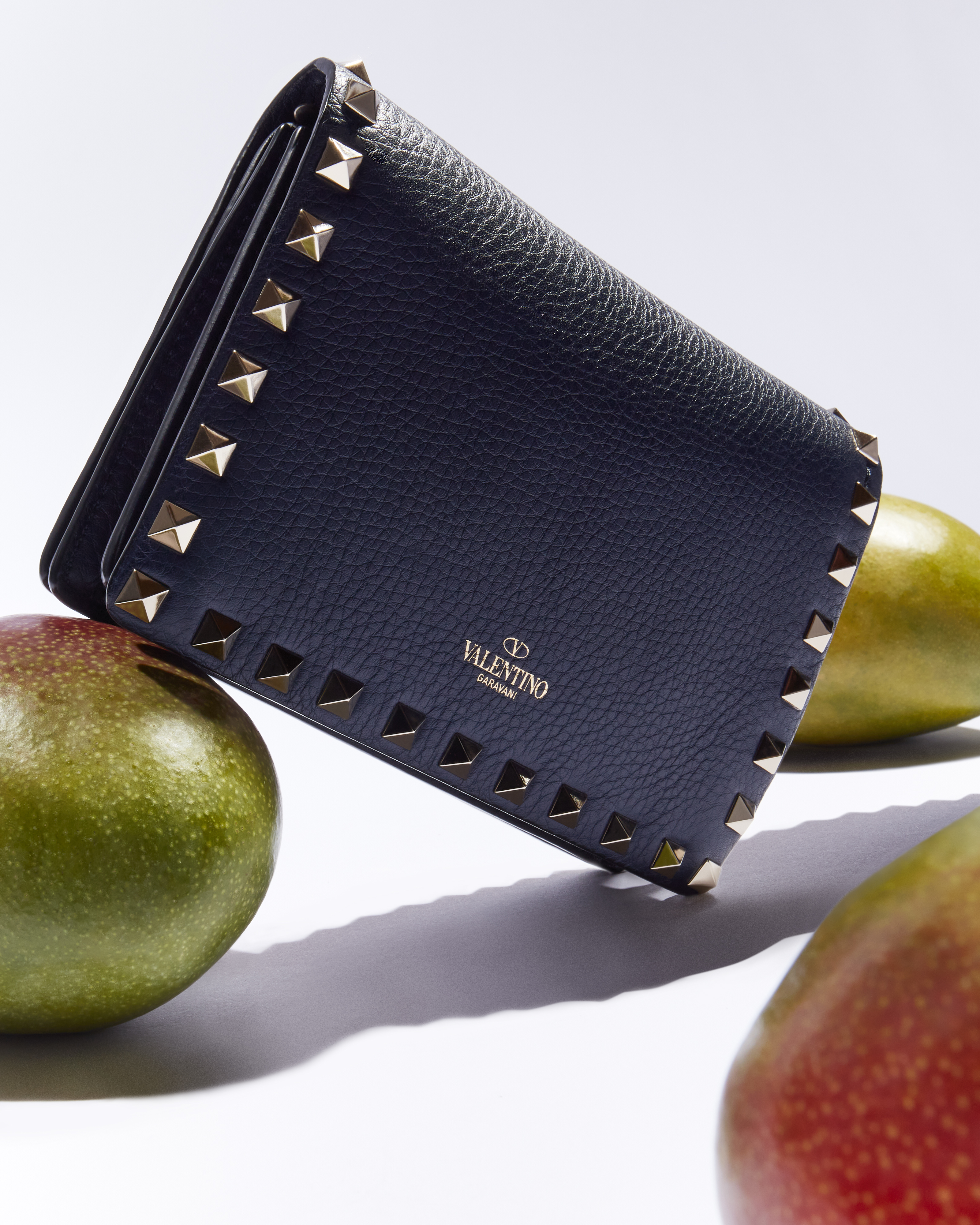 Valentino brand wallet featuring gold studs, contrasted against mango fruit and a bright white background in a studio product shot.