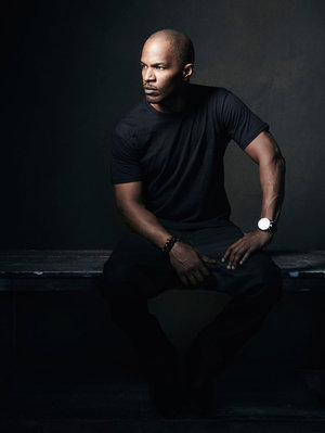 An adult Black man with a bald head poses in an indoor setting, dressed in black attire and accented by a wristwatch and beaded bracelet.