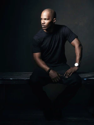 An adult Black man with a bald head poses in an indoor setting, dressed in black attire and accented by a wristwatch and beaded bracelet.