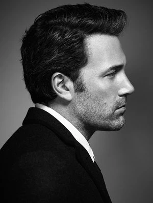 Portrait of a handsome man in a dark coat, captured in a black-and-white studio shot with a grey backdrop and side profile.