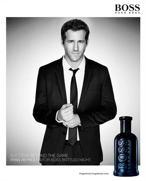 Ryan Reynolds in a black suit and tie, standing in front of a Boss Bottled fragrance bottle in a studio setting.