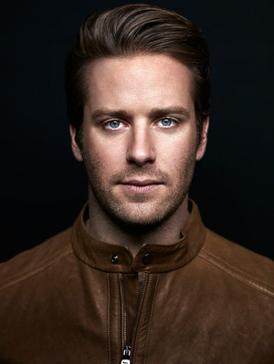Headshot of a blue-eyed male adult with brown hair wearing a brown leather jacket against a dark background in a photo studio.