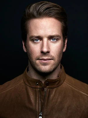 Headshot of a blue-eyed male adult with brown hair wearing a brown leather jacket against a dark background in a photo studio.