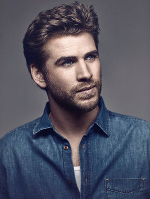 Attractive adult male portrait: Brunette hair, blue eyes, denim shirt, light beard, grey backdrop, neutral expression, looking off-camera.