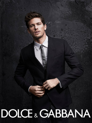 Attractive man poses indoors wearing a black suit with a white shirt and dark tie, hands near his jacket button, against a dark background.