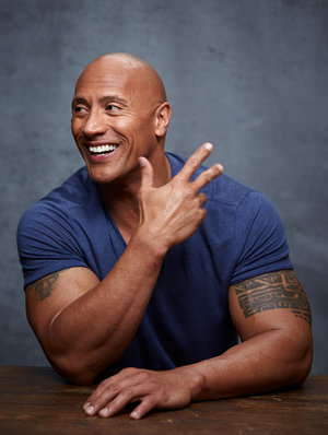 Portrait of Dwayne Johnson smiling, wearing a blue t-shirt, posing at a wooden table with a textured grey background.