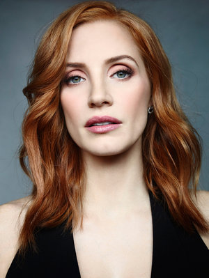 Close-up portrait of a redhead woman with fair skin, wearing a black top, gazing at the camera against a muted blue background.