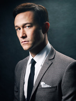 Headshot of Joseph Gordon-Levitt in a grey suit, white shirt, and black tie, set against a dark, moody background in portrait orientation.