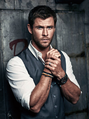 Indoor portrait of a male adult with short brown hair, wearing a grey vest, white shirt, watch, and bracelets, standing against a grey metal wall.