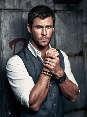 Indoor portrait of a male adult with short brown hair, wearing a grey vest, white shirt, watch, and bracelets, standing against a grey metal wall.