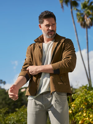 Man in a brown jacket poses outdoors with palm trees and blue sky in the background. He is wearing a light-colored shirt and pants.