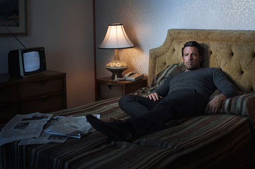 Man in grey sweater and dark trousers reclines on a striped bed in a dimly lit room with an old television set on dresser.