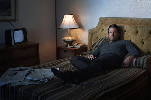 Man in grey sweater and dark trousers reclines on a striped bed in a dimly lit room with an old television set on dresser.