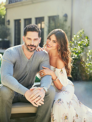 Couple seated outdoors in daylight: a bearded man in a gray sweater and a woman in a floral dress smile for the camera.