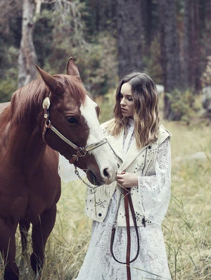 Woman in lace dress and leather vest stands beside a brown and white horse in a wooded field with tall grass and trees.
