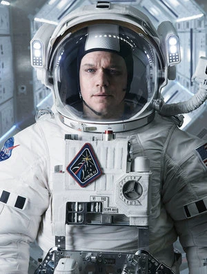 Close-up shot of a serious man in a white astronaut suit with helmet and NASA patch against a gray spacecraft interior backdrop.