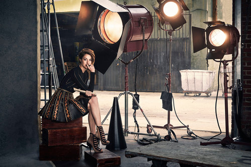 A woman in a black top and patterned skirt sits on wooden boxes in a photo studio surrounded by lighting equipment.