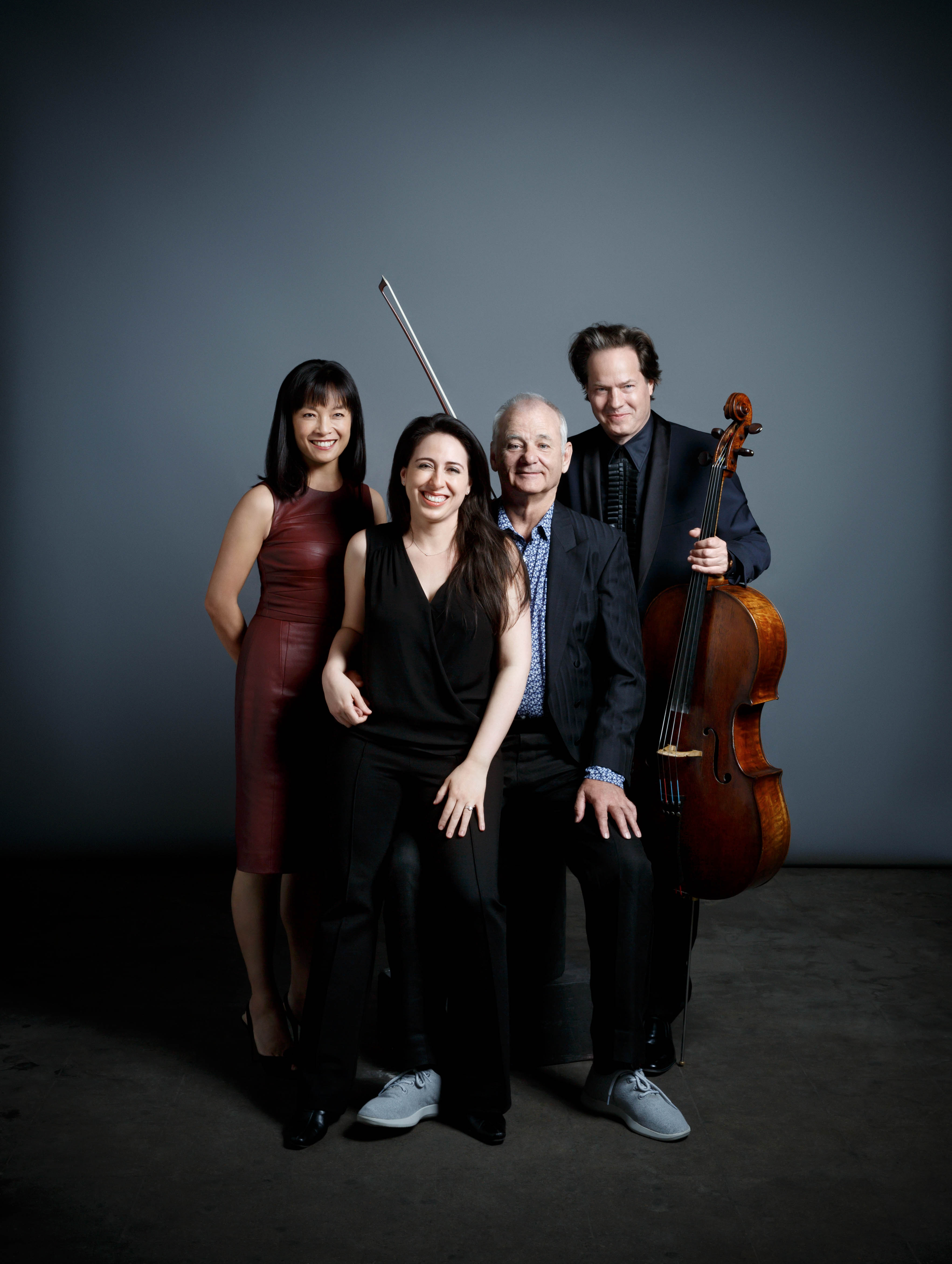 Group studio portrait of two women and two men including Bill Murray with cello against a neutral gray backdrop. Formal attire.