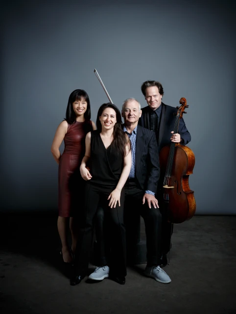 Group studio portrait of two women and two men including Bill Murray with cello against a neutral gray backdrop. Formal attire.